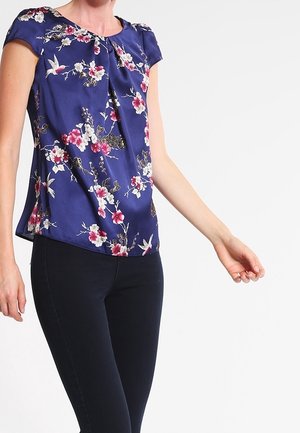 Donna che indossa una camicetta blu navy a maniche corte con motivo floreale e di uccelli rosa, abbinata a pantaloni neri aderenti su sfondo bianco.