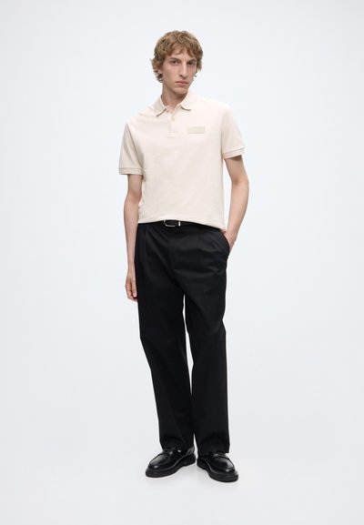 Jeune homme debout, main dans la poche, portant un polo couleur crème, un pantalon noir et des chaussures noires sur fond blanc uni.