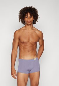 Lila Baumwoll-Boxershorts mit einer weichen Textur, einem breiten Bund und einem kleinen Etikett auf der linken Vorderseite. Modell steht aufrecht.