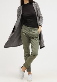 Svart ribbad skjorta, olivgröna byxor och en lång svart-och-vit mönstrad kofta, kombinerat med vita sneakers, stående framför en neutral bakgrund.
