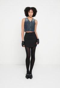 Krótkia kamizelka jeansowa z przednim zamkiem błyskawicznym, zestawiona z czarną mini spódnicą o okrągłym brzegu, czarnymi rajstopami i czarnymi butami z ostrym noskiem.