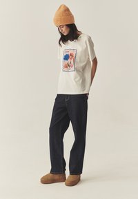 Biała koszulka z kwiatowym wzorem w kolorach niebieskim i różowym, zestawiona z ciemnoniebieskimi jeansami i jasnobrązowymi butami wsuwanymi. Modelka nosi pomarańczową, prążkowaną czapkę beanie.