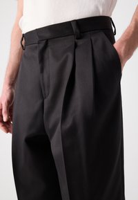 Schwarze, gefaltete Hosen mit glatter Textur, mit Seitentaschen und einem stabilen Bündchen. Nahaufnahme, die die Designdetails hervorhebt.