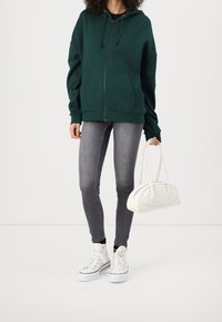 Sudadera verde con capucha y cremallera frontal, combinada con jeans ajustados grises y zapatillas blancas. Bolso blanco con diseño estructurado.