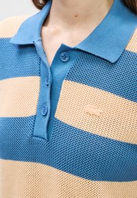 Κοντινό πλάνο από μπλε και μπεζ ριγέ πλεκτό πουκάμισο πόλο με τρία κουμπιά και γιακά, φορεμένο από ένα άτομο.