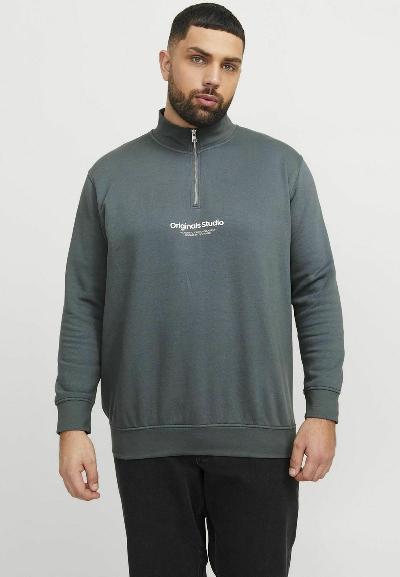 Sudadera gris con cremallera, de manga larga, con puños acanalados y un logotipo impreso en blanco en el pecho. La tela parece suave y lisa.