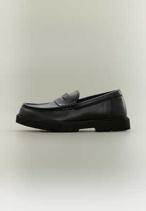 Mocassin en cuir noir avec une semelle en caoutchouc épaisse, un petit talon et une bride décorative à l’avant.