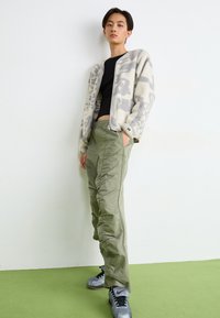 Witte fleece cardigan met grijs abstract patroon, gecombineerd met olijfgroene cargo pants met zijzakken en geplooide details; grijze sneakers.