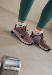 Zapatillas deportivas de color rosa claro con parte superior de malla, detalles en negro y paneles laterales notables, combinadas con calcetines blancos que tienen un logo negro.