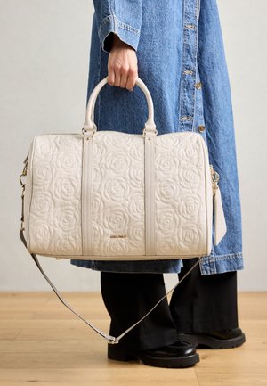 Biely prešívaný kabelka s kvetinovým vzorom, dvojité úchyty a odnímateľný ramenný popruh. Ozdobené zlatými doplnkami, vystavené na džínsovom oblečení.