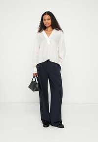 Weiße, langärmlige Bluse mit V-Ausschnitt und plissierten Details, kombiniert mit marineblauen, weit geschnittenen Hosen. Das Model hält eine kleine schwarze Handtasche.