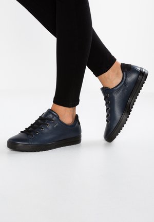 Personne portant des leggings noirs et des chaussures en cuir bleu marine à lacets, avec un pied levé sur un fond de studio blanc.