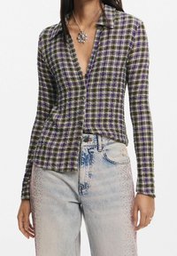 Chemise à boutons en motif écossais vert foncé et violet, avec un col en V profond et des manches longues, assortie à un jean bleu clair avec des embellissements.
