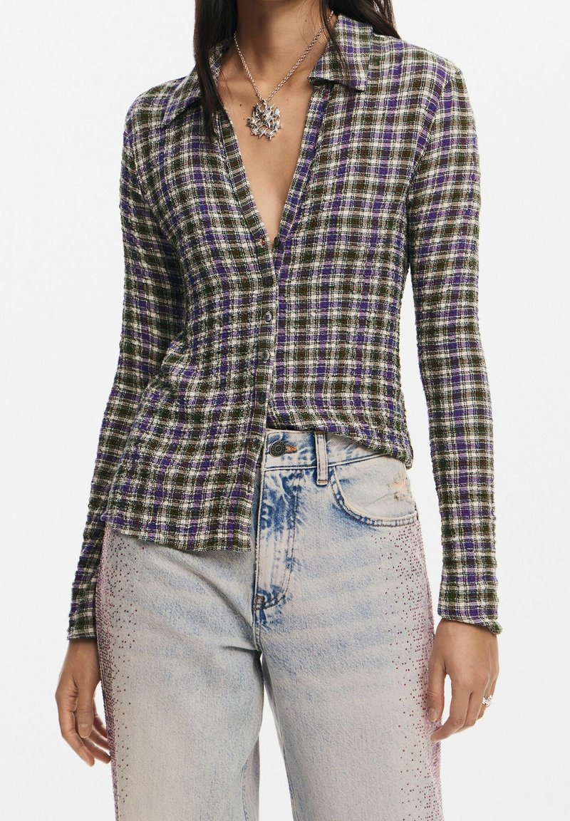 Chemise à boutons en motif écossais vert foncé et violet, avec un col en V profond et des manches longues, assortie à un jean bleu clair avec des embellissements.