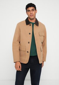 Tan bomuldjakke med fløjlskrage, fire forlommer og knaplukning. Båret over en grøn sweater og mørke bukser.