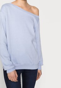 Sudadera de color azul claro con diseño de hombros al aire, puños acanalados y cinturilla, hecha de una tela suave. Llevada con jeans de mezclilla oscura.