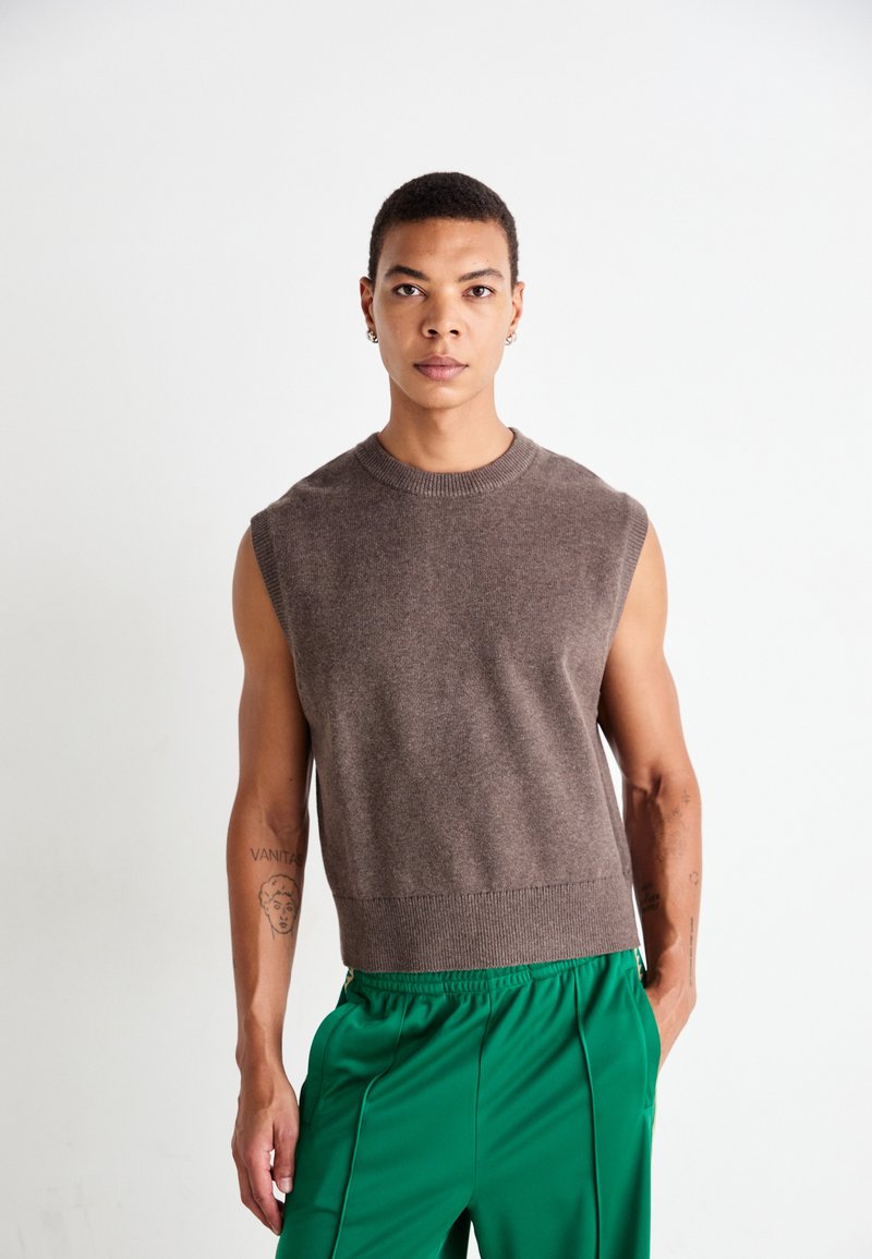 Jeune personne avec les cheveux courts portant un débardeur en tricot marron et un pantalon vert, se tenant contre un fond blanc uni.