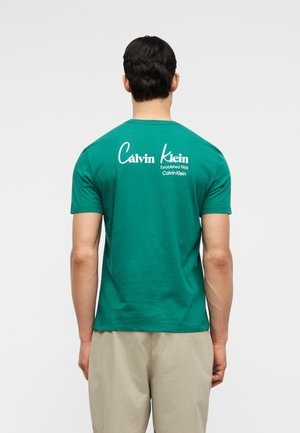 Bărbat purtând un tricou verde Calvin Klein cu logo și text alb pe spate, asortat cu pantaloni bej, stând într-un decor simplu.