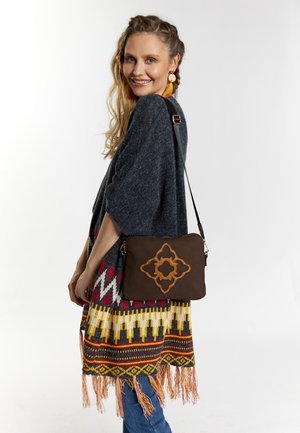 Borsa a tracolla in pelle marrone con motivo ricamato intricato; modello che indossa un poncho grigio con frange colorate e jeans in denim.