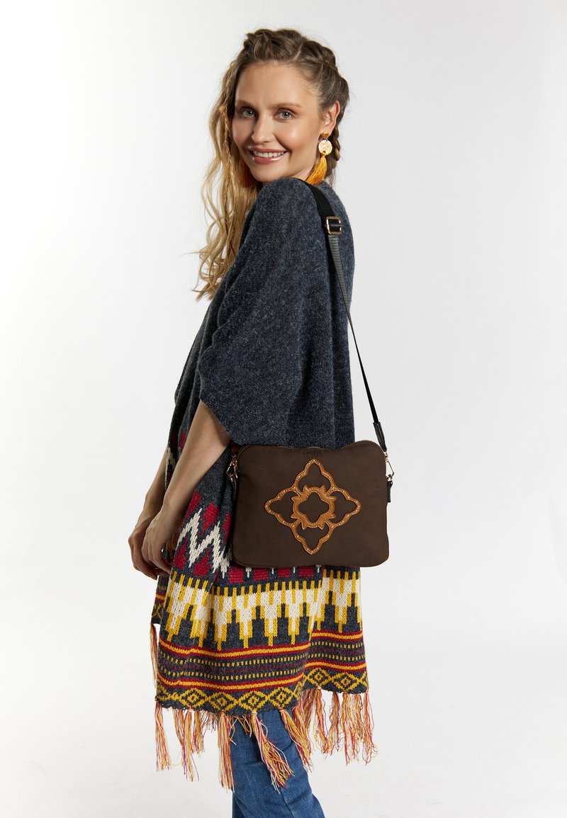 Borsa a tracolla in pelle marrone con motivo ricamato intricato; modello che indossa un poncho grigio con frange colorate e jeans in denim.