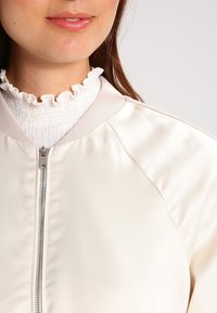 Cream satin bomber jacket featuring a zip closure, ribbed collar, and smooth texture, paired with a white ruffled sweater underneath.