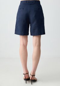 Pantaloni corti in lino blu navy con vestibilità dritta, orli risvoltati e due tasche posteriori. Indossati con sandali con cinturini, che mettono in evidenza un design pulito e minimalista.
