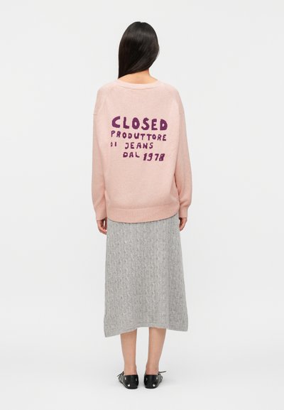 Γυναίκα με μακριά σκούρα μαλλιά που φοράει ροζ πουλόβερ με μοβ γράμματα και γκρι πλεκτή μίντι φούστα, εμφανιζόμενη από πίσω σε λευκό φόντο.