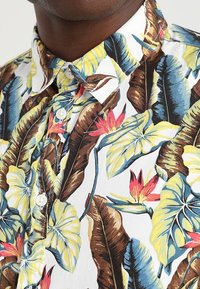 Camicia a maniche corte con pulsanti di colore bianco, decorata con un vivace motivo floreale tropicale, caratterizzato da foglie verdi e fiori rossi. Tessuto di cotone, design con colletto.