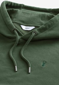 Sweat à capuche vert foncé avec cordons, embouts métalliques, petit logo cerf brodé et étiquette blanche "NEXT" à l'intérieur du col.