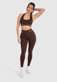 Reggiseno sportivo marrone e leggings a vita alta con scritta bianca "SMIL". Il materiale sembra liscio ed elastico; silhouette aderente senza pattern visibili.
