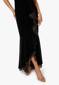 Zwarte fluwelen jurk met een ruffled chiffon overlay en een uitlopende onderkant, gecombineerd met beige sandaaltjes. Soepele textuur en elegant ontwerp.