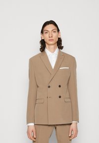 Dvojredni peščeni blazer s klapavicami, dvema sprednjima žepoma in rjavimi gumbi. Ima belo robno rutko. Gladka tekstura.