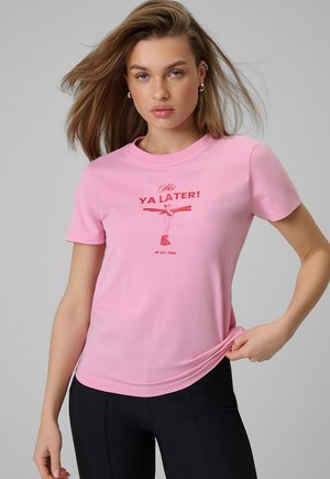 Młoda kobieta z włosami do ramion, ubrana w różowy t-shirt z czerwonym napisem "Ya Later!" oraz czarne spodnie na szarym tle.