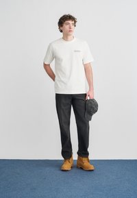 Κοντομάνικη κρεμ μπλούζα με λογότυπο, σκούρο γκρι παντελόνι και τακάκια σε ταμπά χρώμα με κορδόνια. Γκρι καπέλο κρατημένο στο αριστερό χέρι. Απλός σχεδιασμός, casual εφαρμογή.