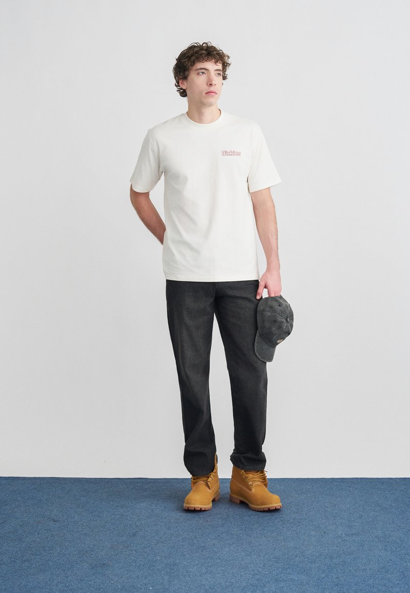 Κοντομάνικη κρεμ μπλούζα με λογότυπο, σκούρο γκρι παντελόνι και τακάκια σε ταμπά χρώμα με κορδόνια. Γκρι καπέλο κρατημένο στο αριστερό χέρι. Απλός σχεδιασμός, casual εφαρμογή.