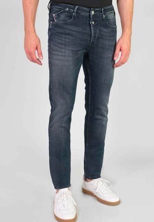 Jean en denim bleu foncé à coupe slim avec cinq poches, léger délavage et texture usée, associés à des baskets blanches.