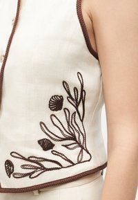 Κοντινή λήψη ενός μπεζ αμάνικου τοπ με καφέ περιθώριο και σκούρα καφέ κεντήματα με μοτίβα θαλάσσιων φυκιών και κοχυλιών στην πλευρά κοντά στη μέση.