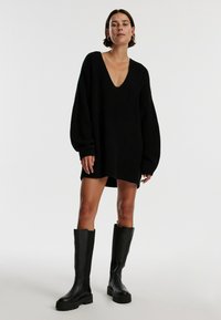 Zwarte gebreide oversized trui jurk met een diepe V-hals, lange ballonmouwen en ribbeltextuur, gecombineerd met zwart kniehoge laarzen.