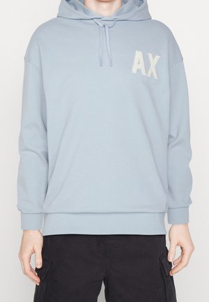 Personne portant un sweat à capuche bleu clair avec les lettres "AX" sur la poitrine et un pantalon cargo noir, debout devant un fond uni.