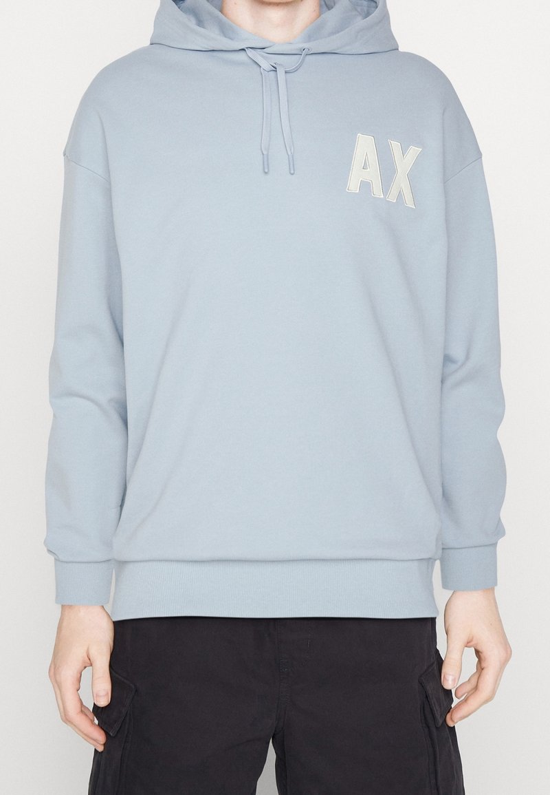 Personne portant un sweat à capuche bleu clair avec les lettres "AX" sur la poitrine et un pantalon cargo noir, debout devant un fond uni.