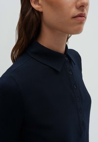 Donna che indossa una camicia blu navy scuro con collo e una patta frontale con bottoni, mostrata dal collo fino a metà torso su uno sfondo semplice.