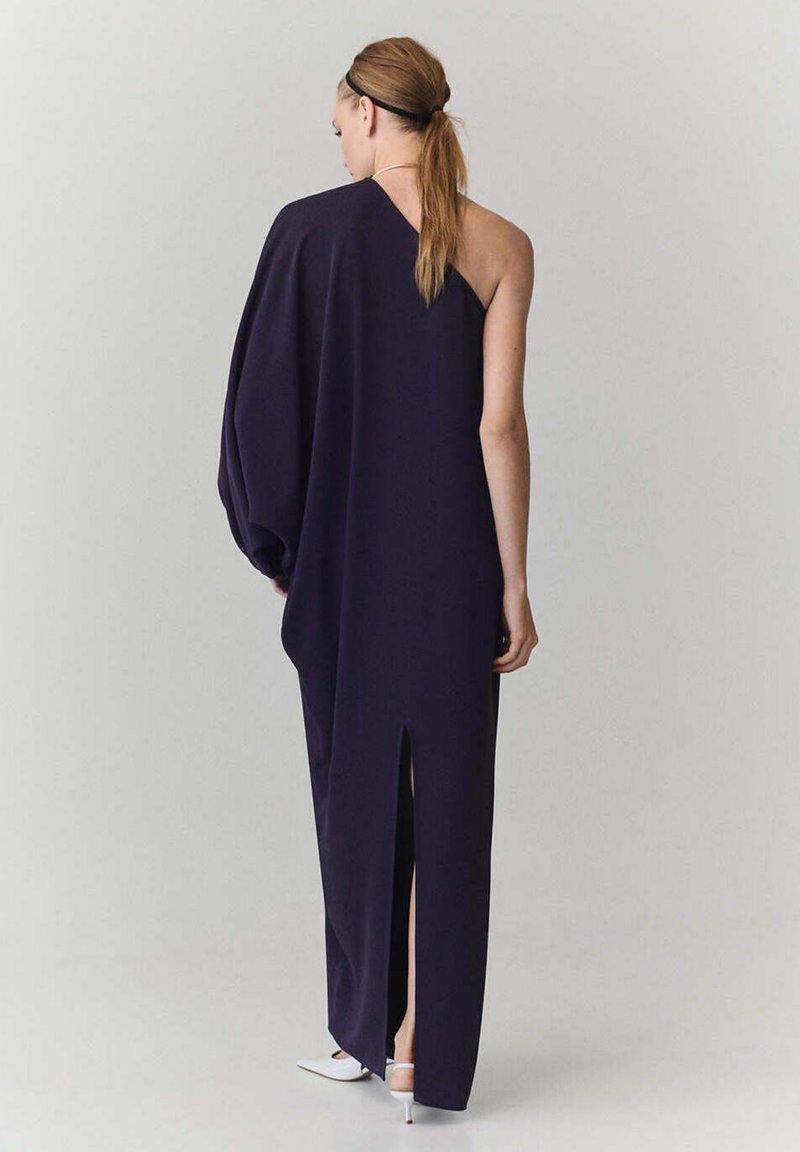Femme avec une queue de cheval portant une longue robe asymétrique violet foncé avec une fente dans le dos et des chaussures blanches à talons hauts sur un fond uni.