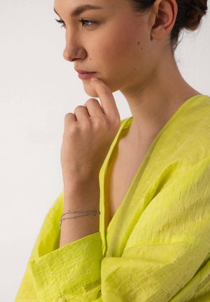 Woman in yellow top resting chin on hand, wearing delicate silver bracelet, looking thoughtfully to the side against a plain background.