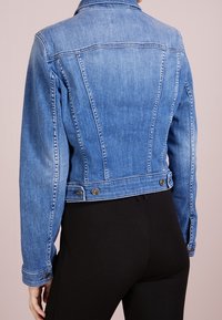 Osoba v zkrácené modré džínové bundě s knoflíkovými detaily a černých úzkých kalhotách, zobrazená zezadu na jednobarevném pozadí.