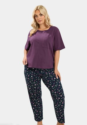 Donna bionda indossa una maglietta viola a maniche corte larga e pantaloni neri con motivi a cuori multicolori, in piedi davanti a uno sfondo semplice.