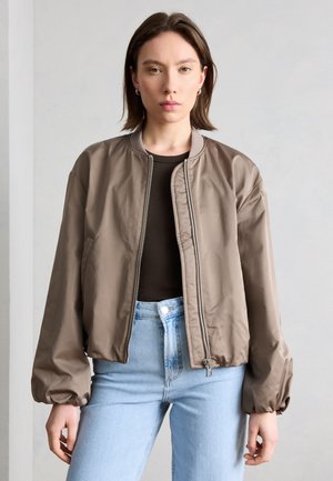 Žena oblečená v taupe bomber bundě přes černý top a světlé modré džíny, stojící před jednobarevným světle šedým pozadím.