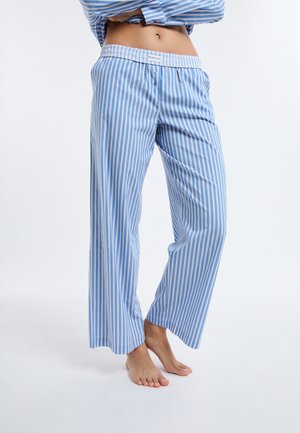 Persona con pijama de rayas verticales azul y blanco, con pantalones y camisa a juego, de pie descalza sobre un fondo blanco liso.