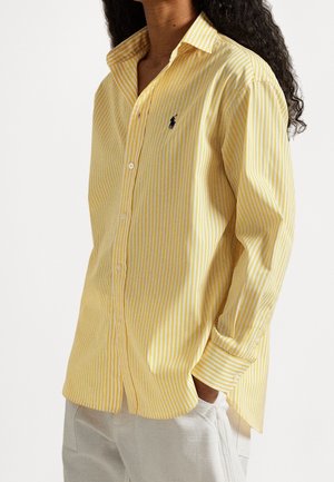 Persoon draagt een geel-wit gestreept overhemd met knopen en een klein zwart polo-logo op de borst en lichte broek met handen in de zakken.