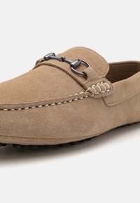 Mocasines de ante beige con puntera redonda, que presentan un acento de cadena redondeada y un pespunte blanco en contraste a lo largo de las costuras. Suela de goma negra.