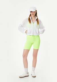 Hvid mesh zip-up jakke over en lys neongrøn bodysuit og matchende cykelshorts. Hvide sneakers og en hvid kasket fuldender looket.